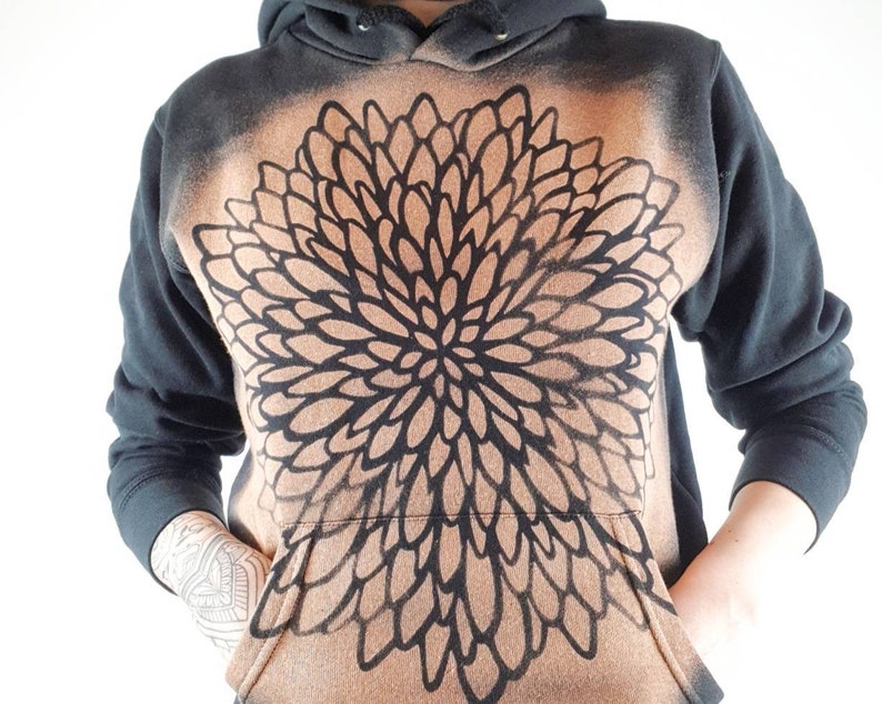 Pu&ograve; includere: Felpa con cappuccio nera con un grande disegno floreale marrone e nero sul davanti. Il disegno &egrave; un motivo a forma di mandala con una forma circolare.