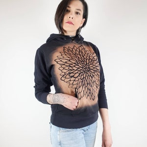 Pu&ograve; includere: Una felpa con cappuccio nera con un disegno di mandala marrone sul davanti. La felpa ha un cappuccio con coulisse e una tasca a marsupio.