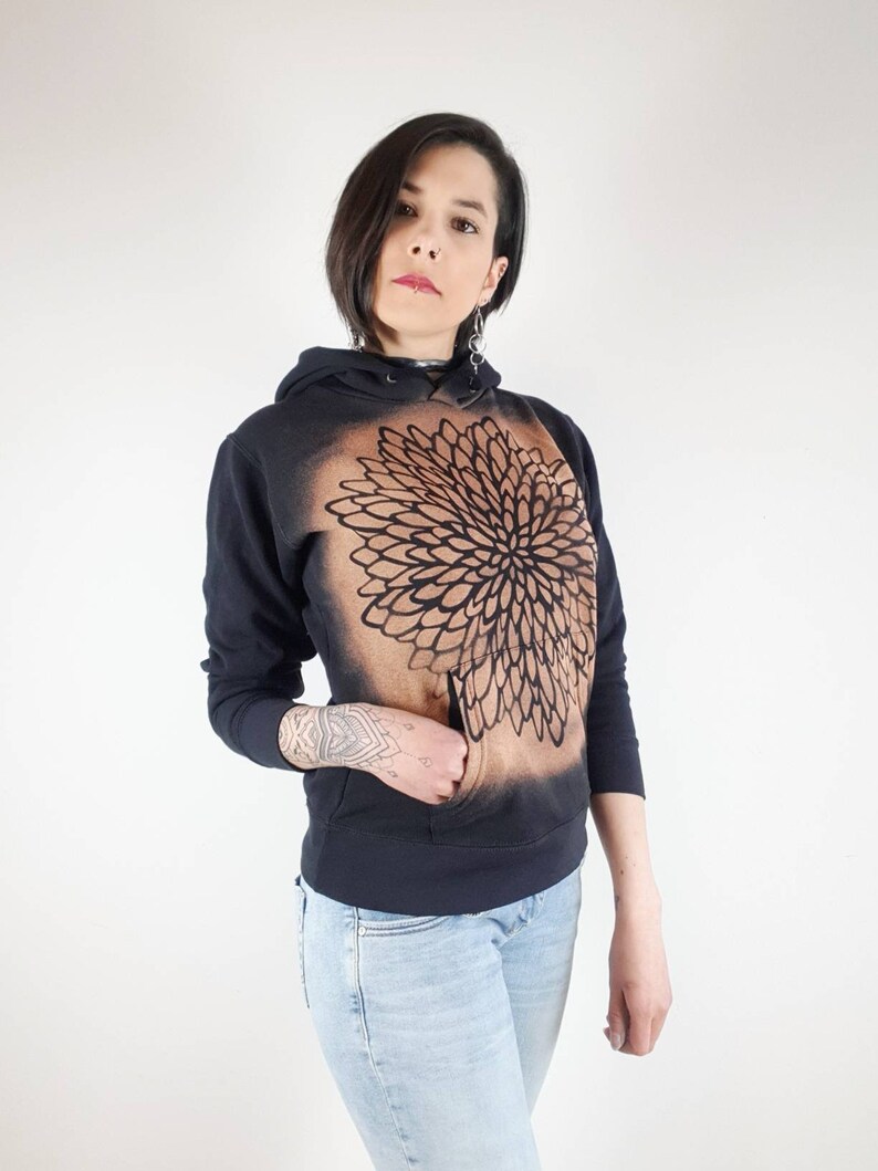 Pu&ograve; includere: Una felpa con cappuccio nera con un disegno di mandala marrone sul davanti. La felpa ha un cappuccio con coulisse e una tasca a marsupio.