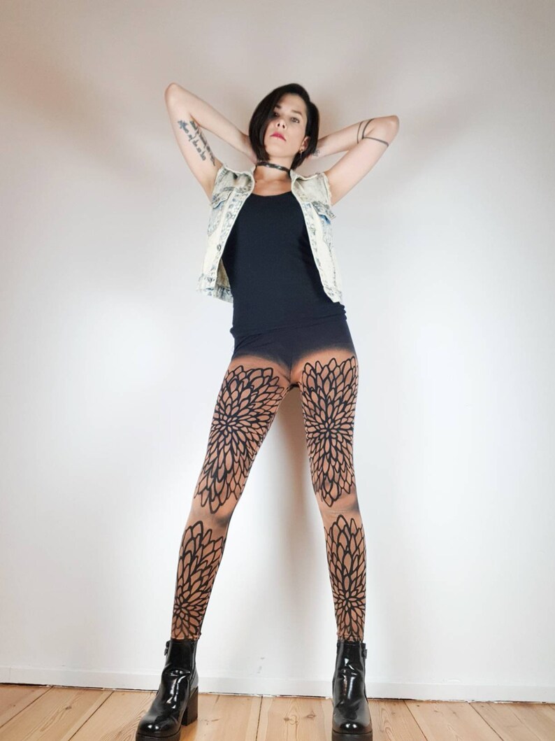 Pu&ograve; includere: Una donna indossa una canotta nera, una giacca di jeans blu chiaro e leggings neri con un motivo floreale. Indossa stivaletti neri alla caviglia e ha i capelli tirati indietro.
