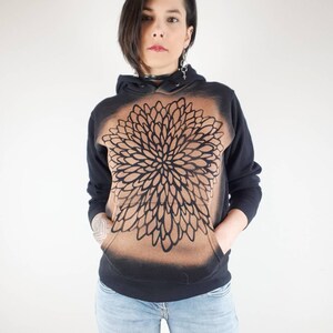 Pu&ograve; includere: Felpa con cappuccio nera con un design a mandala marrone e nero. La felpa ha un cappuccio con coulisse e una tasca a marsupio.