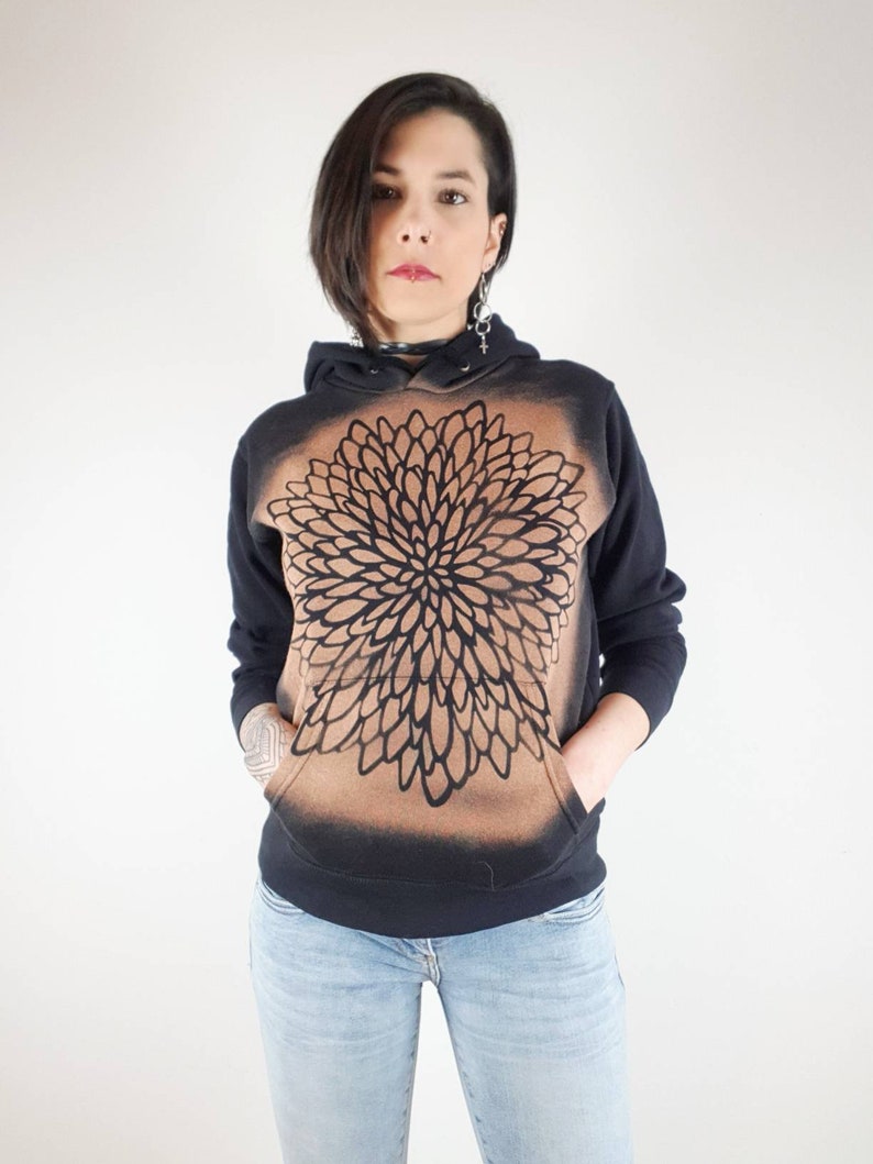 Pu&ograve; includere: Felpa con cappuccio nera con un design a mandala marrone e nero. La felpa ha un cappuccio con coulisse e una tasca a marsupio.
