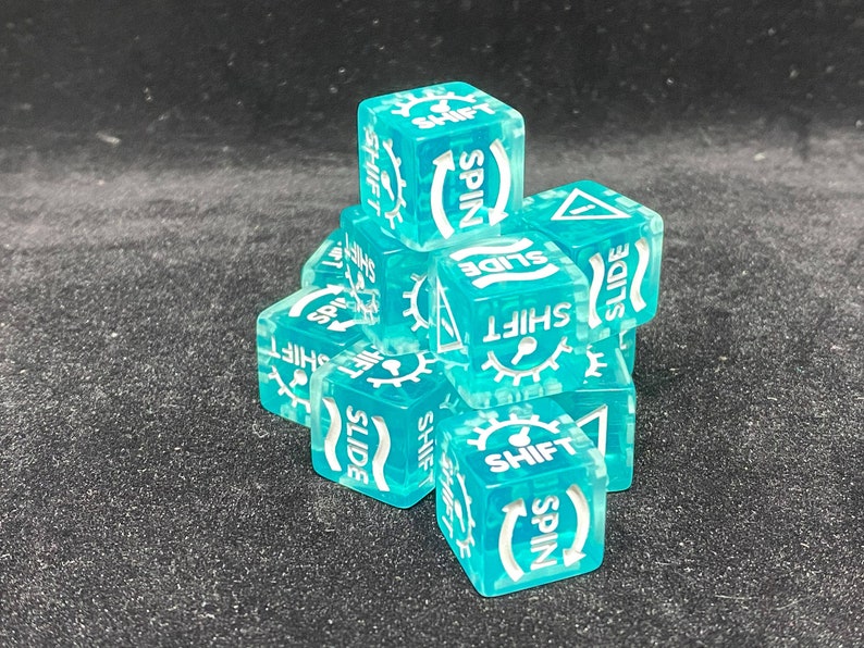 May include: A collection of translucent teal dice with white text and symbols. Each die features the words "SHIFT", "SPIN", and "SLIDE", along with gear and arrow icons. The dice are stacked on a dark surface.