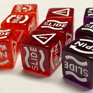 May include: A collection of translucent dice in shades of red and purple. Each die features white text and symbols, including "SLIDE," "SPIN," and "SHIFT." The dice are designed for a board game.