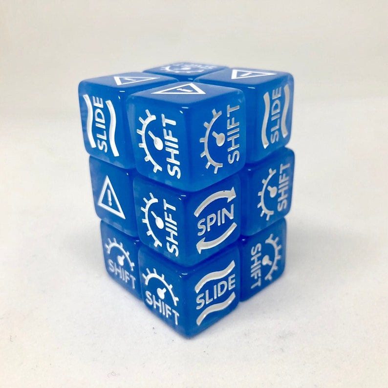 May include: A blue cube puzzle with white text and symbols. The text includes words like "SLIDE", "SHIFT", and "SPIN". The symbols include a gear and an exclamation point inside a triangle. The cube is on a white surface.