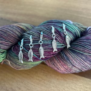 Könnte beinhalten: Fünf silberne, fischförmige Maschenmarkierer werden auf einem Strang mehrfarbigen Garns gezeigt. Das Garn ist eine Mischung aus grünen, violetten und braunen Farbtönen. Die Maschenmarkierer sind an einem Verschluss befestigt, und das Garn liegt auf einer Holzoberfläche.