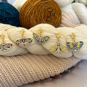 May include: Five gold-toned metal stitch markers shaped like moths, each featuring colourful enamel detailing. The designs include floral patterns, geometric shapes, and star motifs in shades of pink, yellow, black, white, and teal, attached to gold lobster clasps.