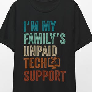 Può includere: T-shirt nera con la scritta "I'M MY FAMILY'S UNPAID TECH SUPPORT" in verde acqua, beige e arancione. Il design include un'icona di monitor di computer e chiave inglese. La maglietta è appesa a una gruccia di legno.