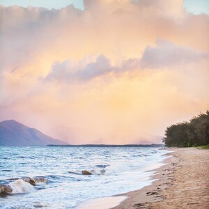 Może przedstawiać: Malowniczy widok piaszczystej plaży z błękitną wodą i białymi falami rozbijającymi się o brzeg. Niebo jest w delikatnym różowym i niebieskim kolorze z puszystymi białymi chmurami. W oddali widać linię zielonych drzew.