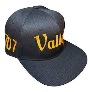 May include: Black baseball cap with a flat brim. The cap features the embroidered text "Valley" in yellow script across the front and "707" on the side, also in yellow. The cap is made of a textured fabric.