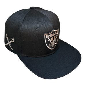 May include: Black baseball cap with a flat brim. The cap features a silver embroidered shield with the text "RN4" and a pirate graphic. Two crossed swords are embroidered on the side.