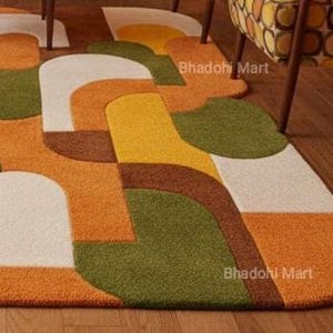 May include: A colorful, geometric area rug with a retro design. The rug features a pattern of curved shapes in shades of orange, green, yellow, brown, and white. The rug is placed on a wooden floor, and the text "Bhadohi Mart" is visible.