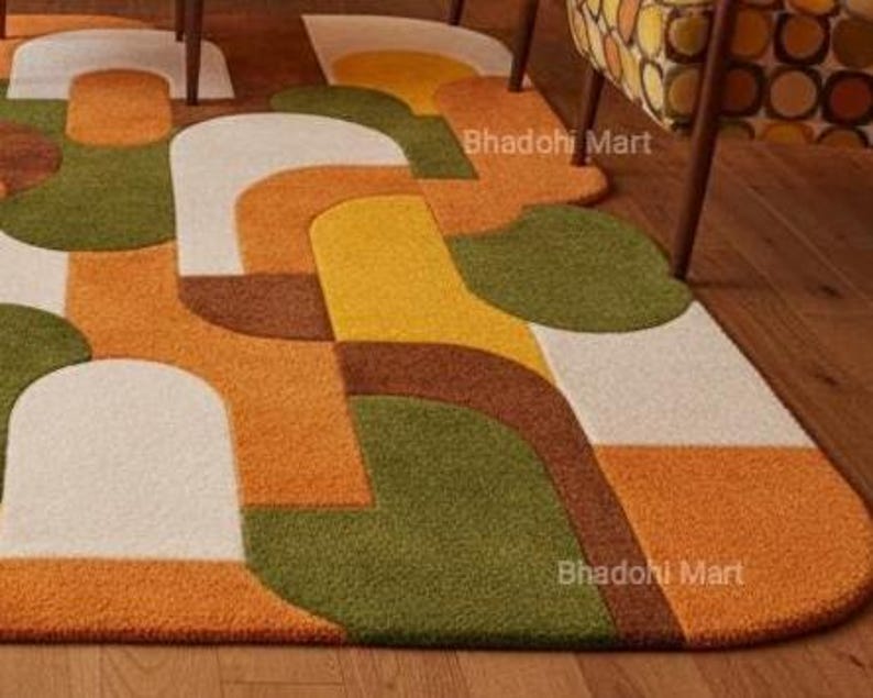 May include: A colorful, geometric area rug with a retro design. The rug features a pattern of curved shapes in shades of orange, green, yellow, brown, and white. The rug is placed on a wooden floor, and the text "Bhadohi Mart" is visible.