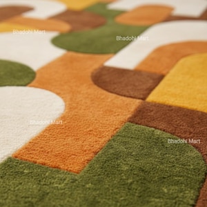 May include: A close-up of a colorful rug with a geometric pattern. The rug features a combination of orange, green, brown, yellow, and white shapes. The text "Bhadohi Mart" is visible in several places on the rug.