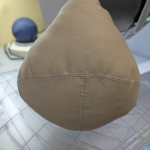 Puede incluir: Un objeto de tela color canela en forma de cúpula con costuras visibles que forman un patrón de cruz en la superficie. El artículo está colocado sobre una superficie de plástico transparente con una regla que muestra medidas en centímetros.