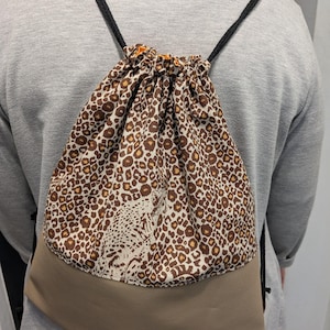Könnte beinhalten: Ein brauner und beiger Rucksack mit Kordelzug mit Leopardenmuster und einer Geparden-Silhouette. Der Rucksack hat schwarze Kordelzüge.