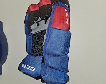 Puede incluir: Un guante de hockey azul y rojo con la marca "CCM" visible. El guante tiene un diseño acolchado con detalles rojos en los dedos y el pulgar. La zona de la muñeca es negra, y el diseño general sugiere un enfoque en la protección y el rendimiento.