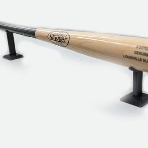 Może przedstawiać: Drewniana pałka do baseballu z czarną rączką i napisem "Louisville Slugger" na boku. Pałka jest wystawiona na czarnym metalowym stojaku.