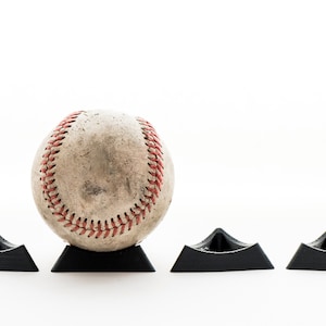 Könnte beinhalten: Ein weißer Baseball mit roten Nähten liegt auf einem schwarzen Baseball-Präsentationsständer. Zwei weitere schwarze Ständer befinden sich auf beiden Seiten des Baseballs.