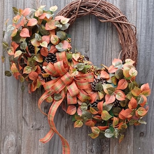 May include: A fall-themed wreath with a grapevine base, adorned with orange, green, and brown leaves, pinecones, and berries. A large plaid ribbon in orange and green adds a decorative touch. The wreath is displayed against a weathered wooden background.
