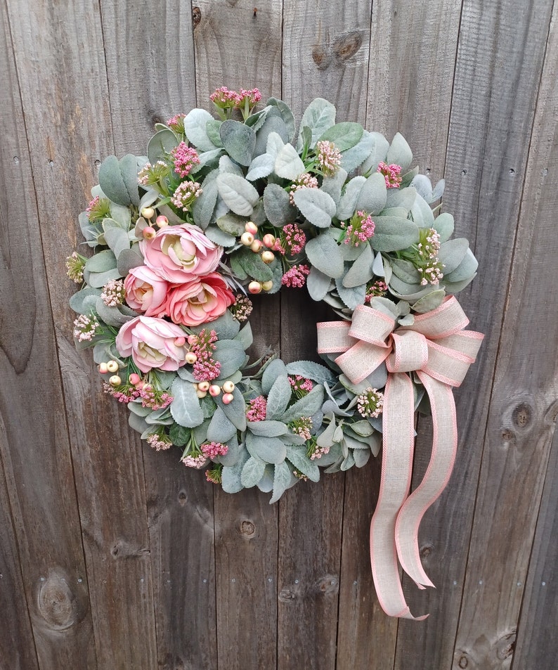 May include: A spring-themed artificial wreath featuring blush pink ranunculus flowers and dusty miller greenery.  A blush pink ribbon adds a touch of elegance. The wreath is approximately 14 inches in diameter and ideal for front door decor, adding a touch of springtime charm to any home.