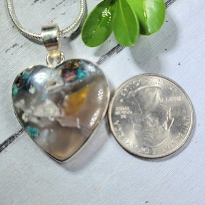 May include: A silver heart-shaped pendant with a clear stone filled with colorful inclusions. The stone is about the size of a US quarter.