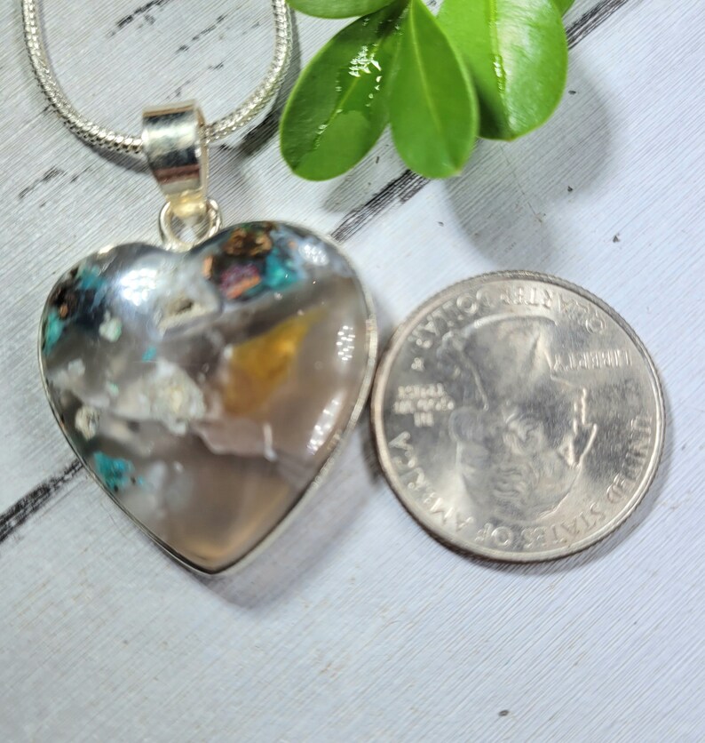 May include: A silver heart-shaped pendant with a clear stone filled with colorful inclusions. The stone is about the size of a US quarter.