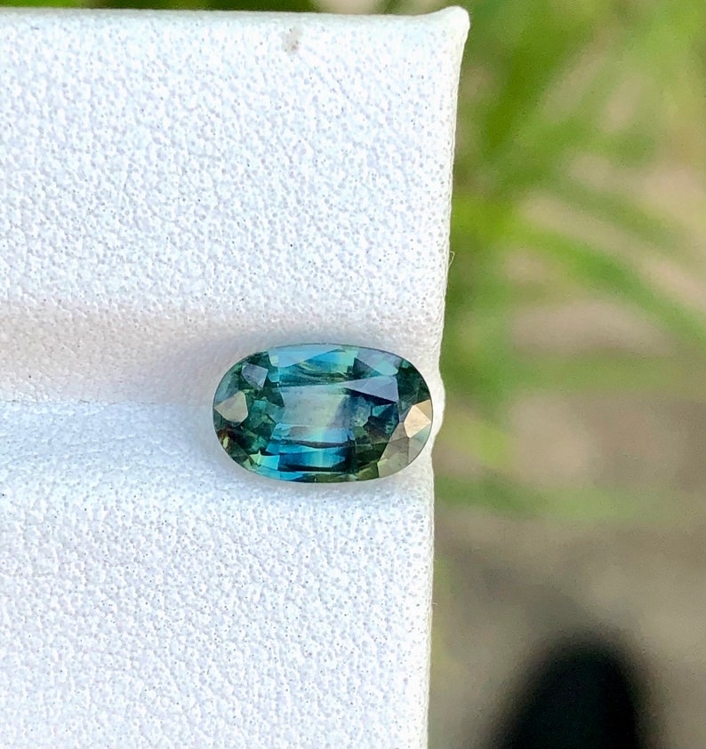 May include: Oval-shaped green gemstone with a blue hue, set on a white background.