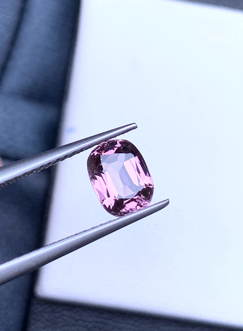 May include: A pink oval-shaped gemstone held by tweezers against a white background.