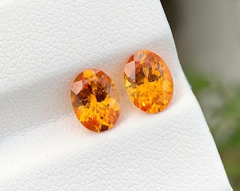 May include: Two oval-shaped orange gemstones on a white background. The gemstones are faceted and have a bright, vibrant color.