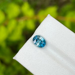 May include: A teal blue oval gemstone with a slight green hue, set on a white background.
