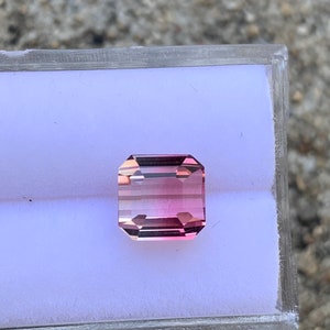 May include: A pink and clear gemstone with a rectangular shape, set in a clear plastic box.