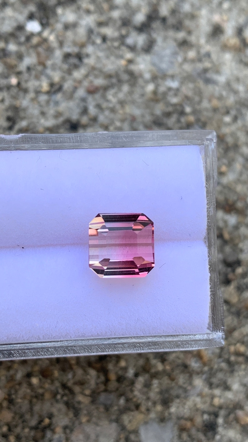 May include: A pink and clear gemstone with a rectangular shape, set in a clear plastic box.