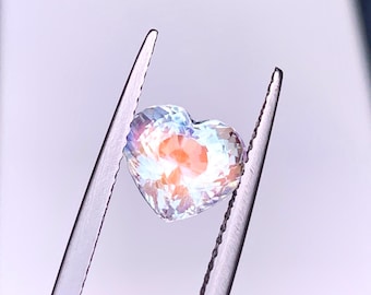 Può includere: Una pietra preziosa a forma di cuore con un effetto arcobaleno iridescente. La pietra preziosa è tenuta in una pinza e sembra essere di colore blu chiaro con un tocco di arancione.