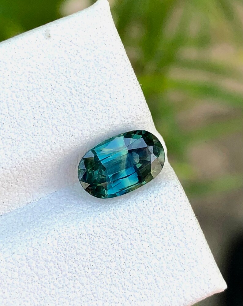 May include: A green oval gemstone with a blue hue, set on a white background.