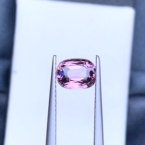 May include: A pink oval-shaped gemstone held in tweezers against a white background.