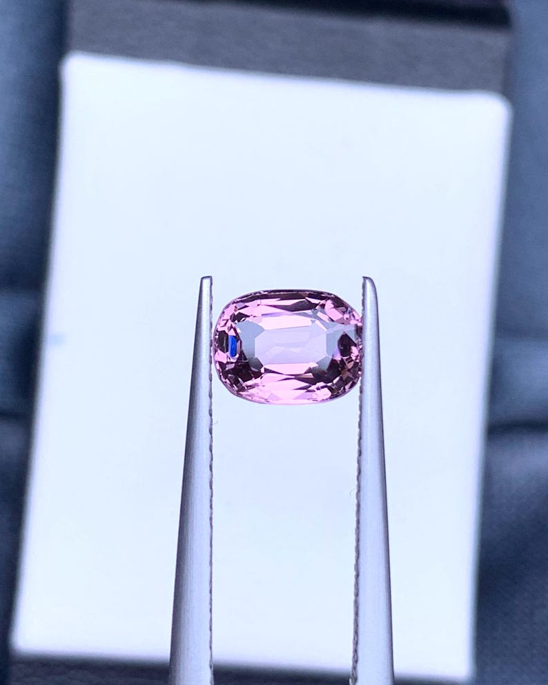 May include: A pink oval-shaped gemstone held in tweezers against a white background.