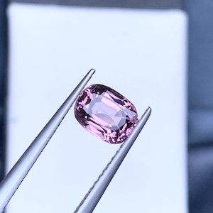 May include: A pink oval-shaped gemstone held by tweezers against a white background.