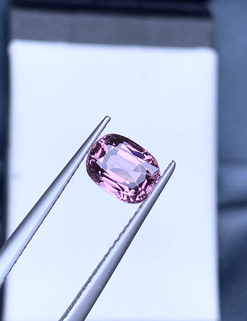 May include: A pink oval-shaped gemstone held by tweezers against a white background.