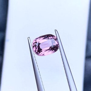 May include: A pink oval-shaped gemstone held by tweezers against a white background.
