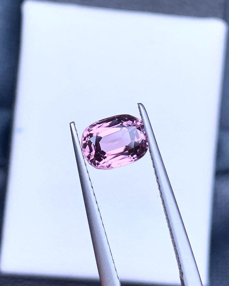 May include: A pink oval-shaped gemstone held by tweezers against a white background.