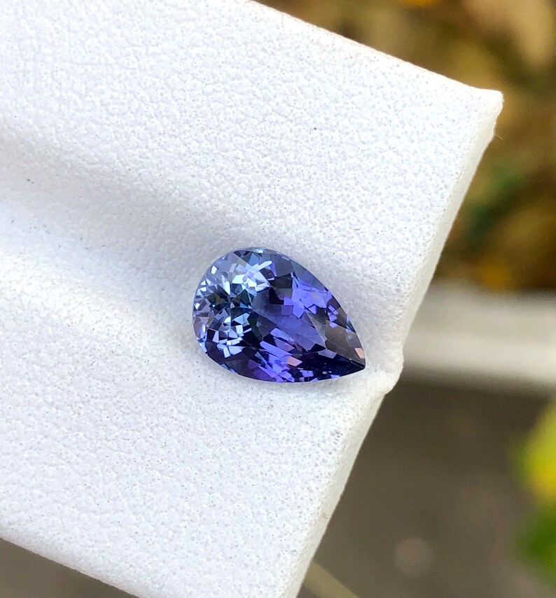 K&ouml;nnte beinhalten: Ein birnenf&ouml;rmiger Edelstein mit einer tiefblauen bis violetten Farbe. Der Edelstein ist auf einem wei&szlig;en Hintergrund platziert.