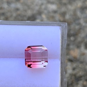 May include: A pink and clear gemstone with a rectangular cut, set in a white display box.