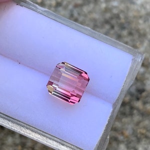 May include: A pink and yellow gemstone with a rectangular shape, set in a white display box.