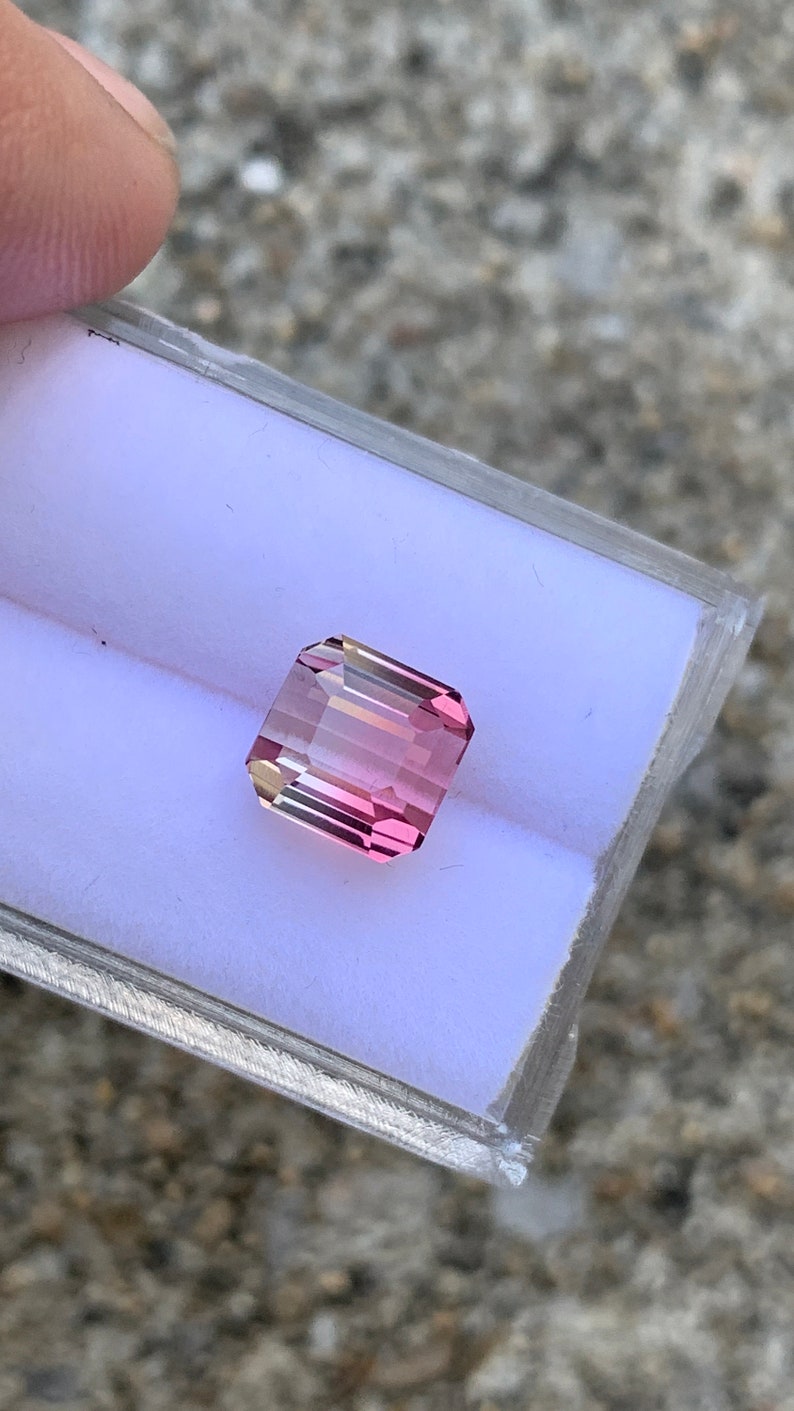 May include: A pink and yellow gemstone with a rectangular shape, set in a white display box.