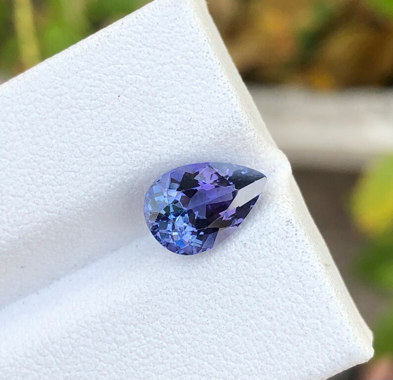 K&ouml;nnte beinhalten: Ein birnenf&ouml;rmiger Edelstein mit einem tiefen violetten Farbton. Der Edelstein ist auf einem wei&szlig;en Pr&auml;sentationsst&auml;nder platziert.