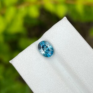 May include: A blue oval gemstone with a slight green hue, displayed on a white surface.