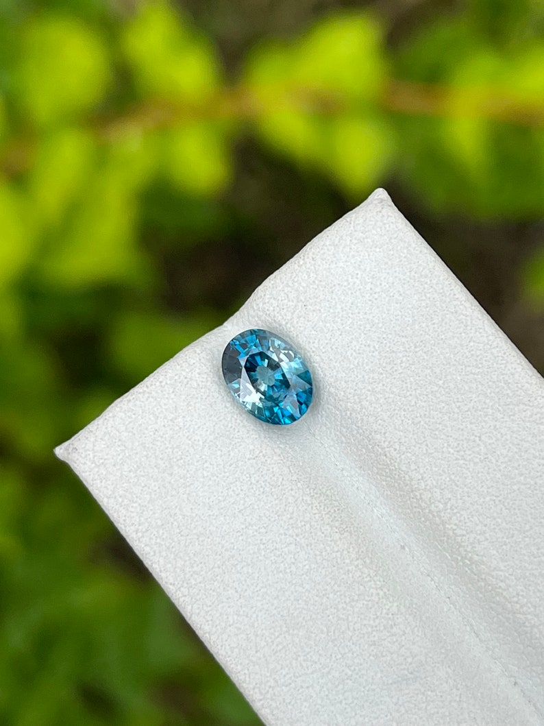 May include: A blue oval gemstone with a slight green hue, displayed on a white surface.