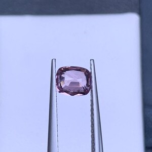 May include: A light pink gemstone with a cushion cut, held in tweezers against a white background.