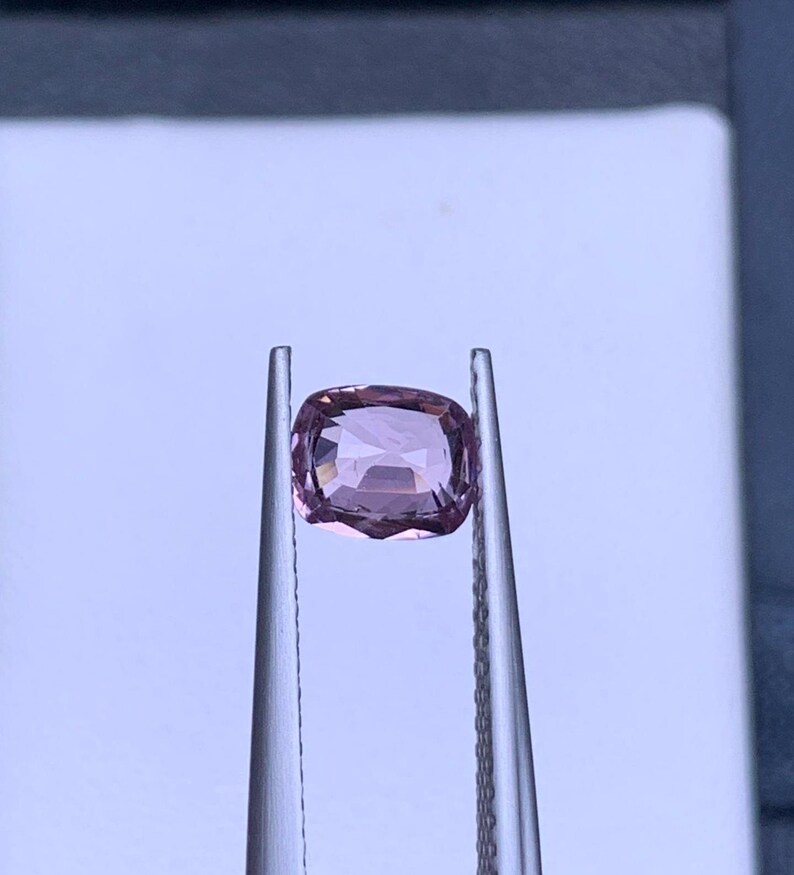 May include: A light pink gemstone with a cushion cut, held in tweezers against a white background.